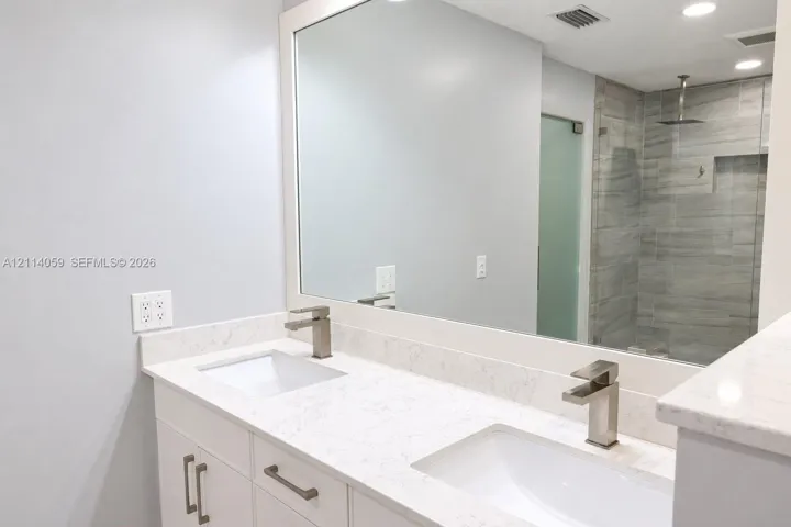 Double vanity featuring white cabinetry, stone-finish countertops, rectangular undermount sinks, and brushed-finish faucets, paired with a walk-in shower encased in glass with tile wall surfacing and a built-in recessed shelf