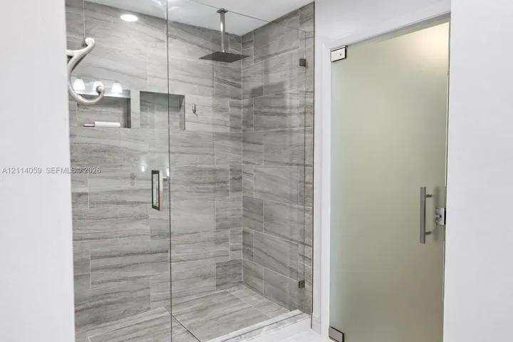 Walk-in shower featuring floor-to-ceiling stone-pattern tile, a ceiling-mounted rainfall showerhead, recessed shelving, and a frameless glass enclosure with a frosted glass entry door