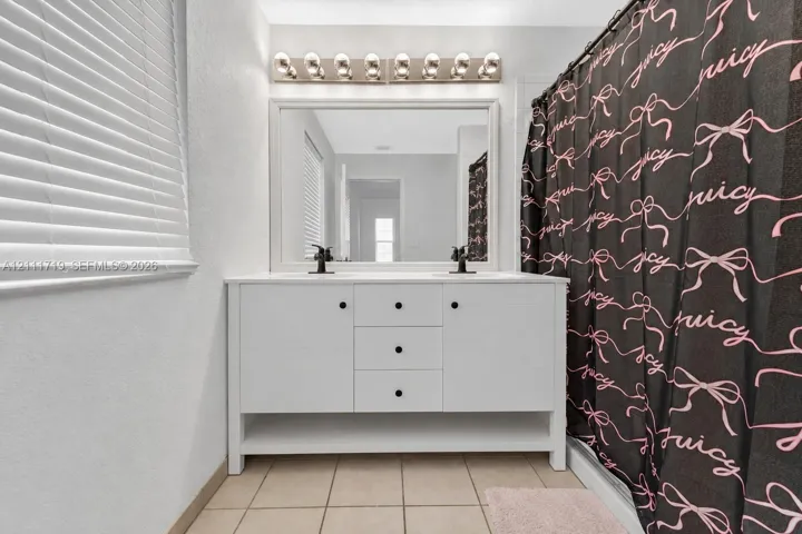 White dual-sink vanity with central drawer storage, large framed mirror, multi-bulb wall-mounted lighting, and ceramic tile flooring