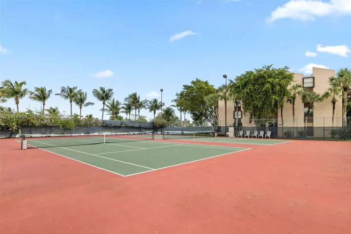 Hard-court tennis facility featuring perimeter chain-link fencing, dual courts, overhead lighting, and mature palm landscaping