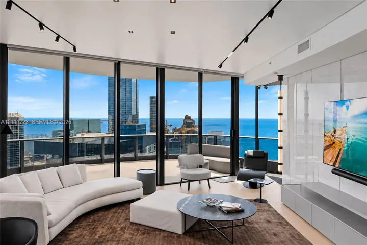 Floor-to-ceiling windows featuring panoramic water and city scenery, expansive curved balcony, stone-finish accent wall with integrated media cabinetry, and recessed track lighting