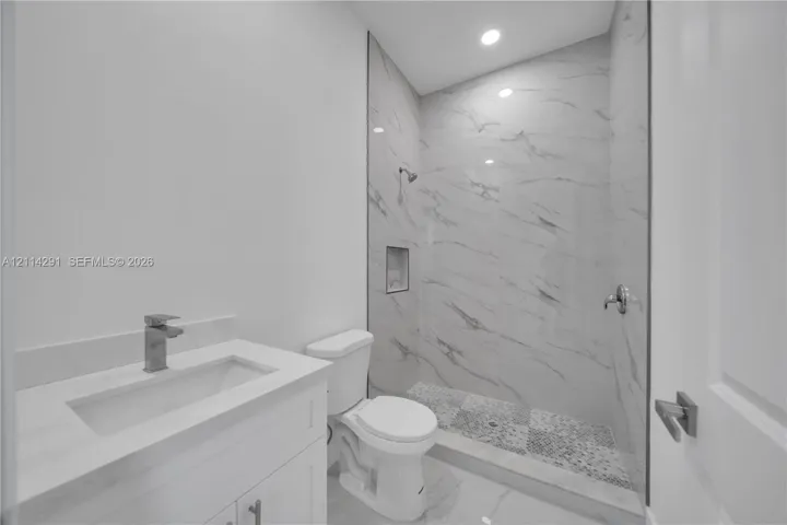 Bathroom featuring a white vanity with integrated rectangular sink and stone-finish countertop, shower stall with floor-to-ceiling stone-finish tile surround and integrated wall niche, pebble-texture shower floor, and recessed ceiling lighting