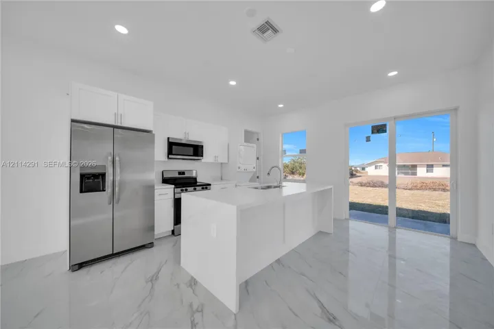Kitchen featuring white shaker-style cabinetry, stainless steel appliances, a waterfall-edge stone island with a stainless steel undermount sink, recessed lighting, and large-format porcelain tile flooring
