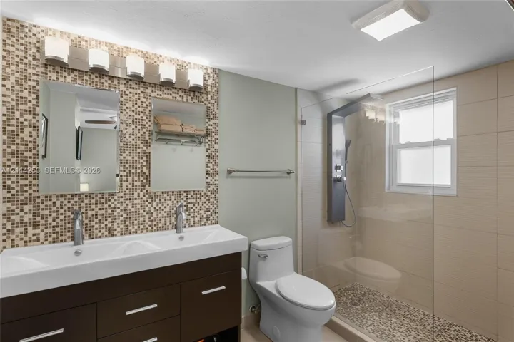 Bathroom featuring a dual-basin vanity with a dark finish, mosaic tile backsplash, frameless glass shower enclosure, integrated shower panel system, and pebble-style shower floor tile