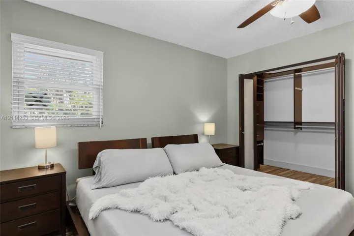 Neutral-toned room featuring a ceiling fan with integrated lighting, horizontal window blinds, and a built-in closet system with shelving and hanging rods