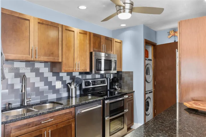 Kitchen featuring wood cabinetry, stone-finish countertops, geometric tile backsplash, stainless steel appliances, and integrated laundry closet
