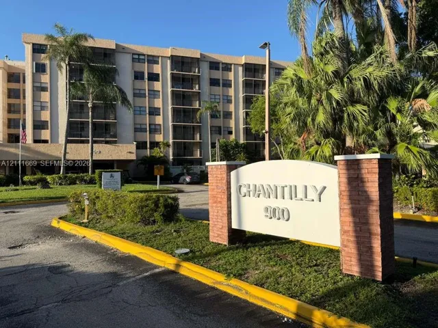 Multi-story residential building featuring covered balconies, brick-accented monument signage, mature palm trees, and manicured landscaping