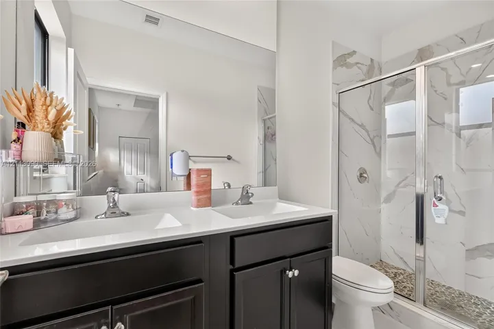 Double vanity with dark cabinetry, white countertops, dual undermount sinks, and a walk-in shower featuring tile surround and a glass door