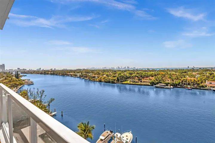 Balcony featuring a white metal railing overlooking a wide waterway, coastal vegetation, and a distant city skyline
