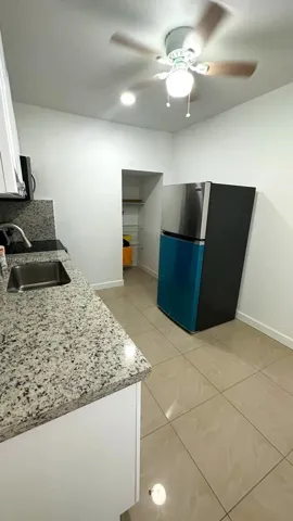 Kitchen featuring stone-finish countertops, stainless steel undermount sink, large-format tile flooring, ceiling fan with integrated lighting, and reach-in storage pantry