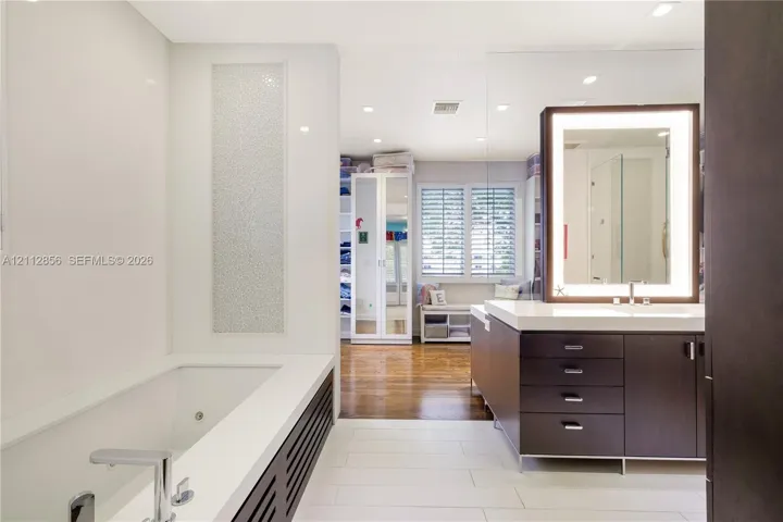 Contemporary bathroom featuring a deep-soaking tub with a tiled surround, custom dark-finish vanity cabinetry with a stone countertop, an integrated LED-lighted mirror, and large-format porcelain floor tile