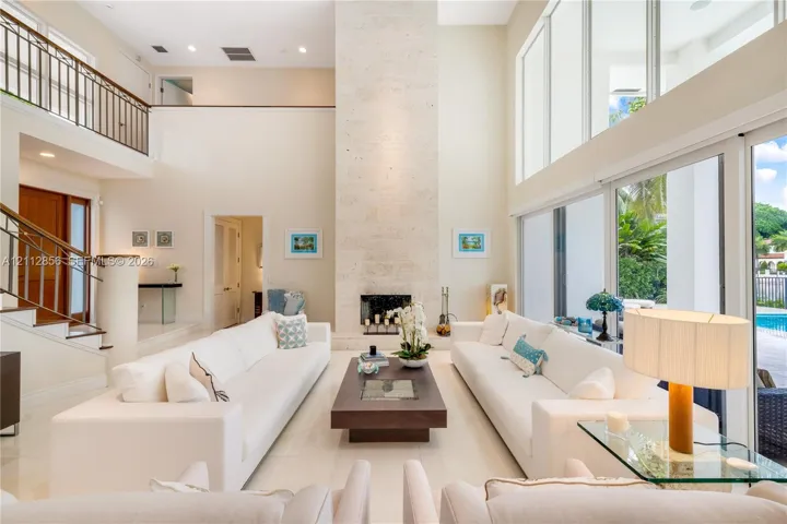 Double-height living area featuring a floor-to-ceiling stone fireplace, expansive glass wall panels, a mezzanine balcony with metal railing, and polished stone flooring