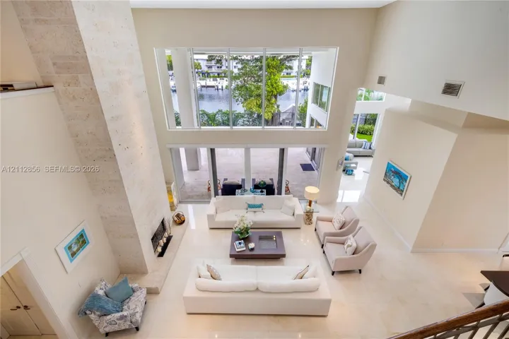 Double-height living area with natural stone fireplace surround, floor-to-ceiling windows, light-toned tile flooring, and direct access to an outdoor patio