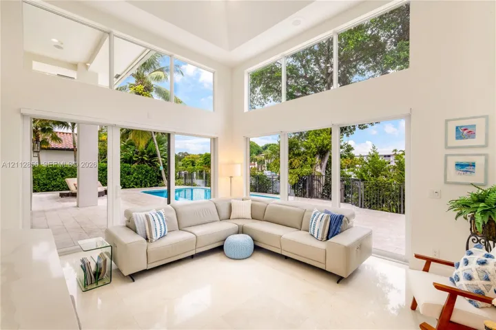 Double-height ceilings, floor-to-ceiling windows, polished stone flooring, and multiple sliding glass doors opening to a paved patio with a swimming pool