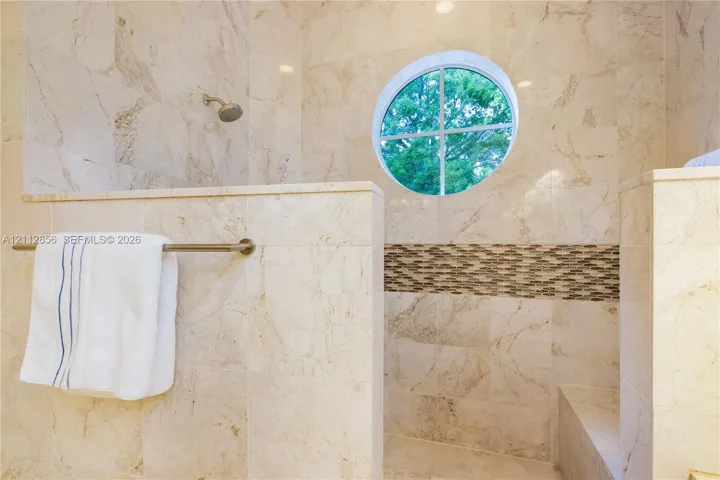 Walk-in shower featuring floor-to-ceiling stone tile, circular accent window, horizontal decorative stone inlay, built-in tiled bench, and wall-mounted shower head