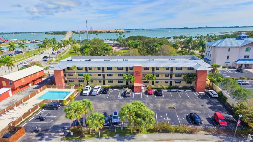 Multi-story residential building featuring a metal roof, dedicated paved parking area, ground-level outdoor swimming pool, and tropical landscaping