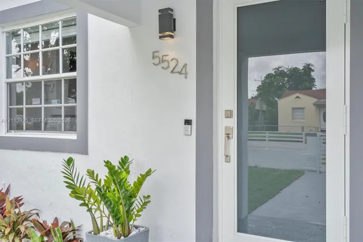 Exterior entryway featuring a white-framed glass door, multi-pane window with dark trim, wall-mounted rectangular outdoor light fixture, and metallic house numerals