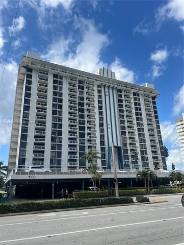 High-rise residential exterior featuring multiple tiers of balconies with metal railings, structural blue-toned architectural accents, and a covered ground-level parking area