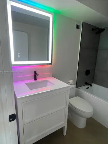 White cabinetry vanity with a solid-surface countertop and undermount sink, integrated matte black faucet, wall-mounted LED-backlit mirror, and tub enclosure featuring large-format dark tiling with matte black hardware
