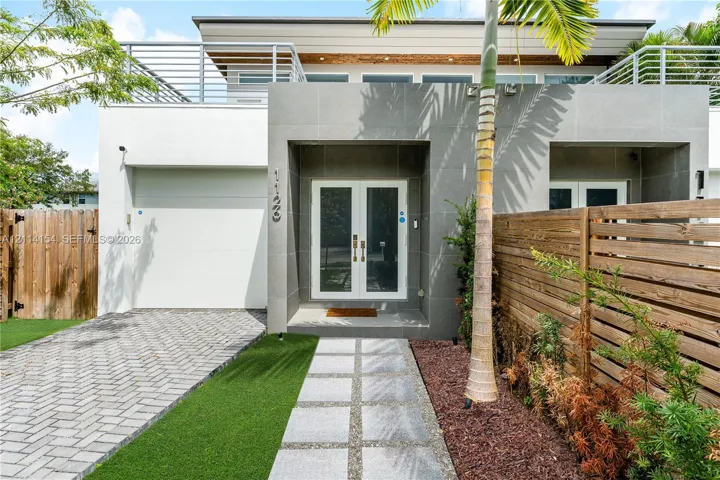 Modern exterior featuring a paved stone driveway, synthetic turf lawn, second-story balcony with metal railings, and a symmetrical glass-panel double door entrance