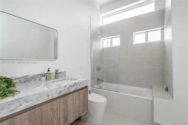 Floating wood-finish vanity with stone-surface countertop, rectangular undermount sink, glass-enclosed tub shower with full-height tile surround, and clerestory window