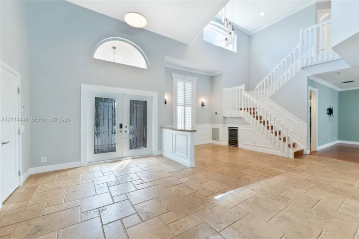 Double-height entryway featuring large-format tile flooring, a grand staircase with decorative balustrades, double entry doors with glass inserts, an arched transom window, and built-in under-stair cabinetry