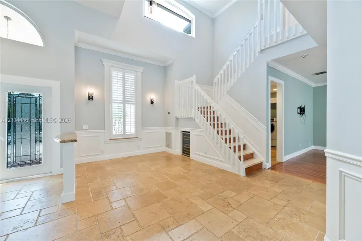Grand foyer featuring vaulted ceilings, natural stone tile flooring, a staircase with custom white millwork, decorative wall paneling, and an integrated wine chiller