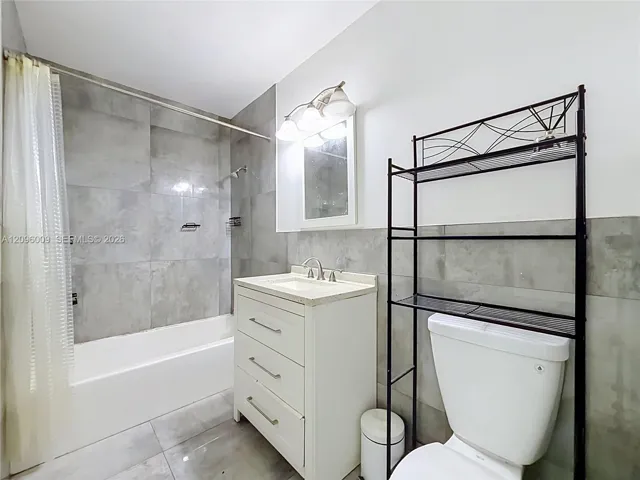 Bathroom featuring large-format grey tile surround, white vanity cabinet with stone-finish countertop, undermount sink, and three-light wall fixture