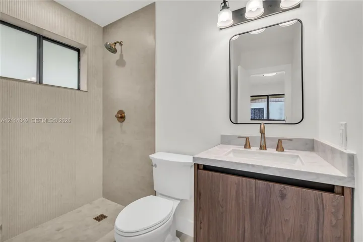 Bathroom featuring a wood-finish vanity with a stone countertop, wall-mounted rectangular mirror, three-light fixture, and a walk-in shower with large-format tile surround