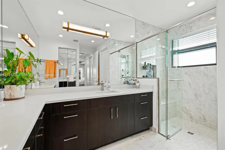 Bathroom featuring a dual-sink dark wood vanity with white countertops, a full-width mirror, recessed lighting, and a glass-enclosed shower with stone-tile surround