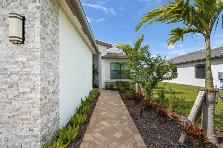 Stucco exterior featuring stacked stone veneer accents, paver walkway, wall-mounted outdoor lighting, and landscaped beds with palm trees
