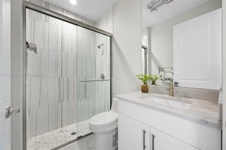 Bathroom featuring a walk-in glass shower with vertical-patterned wall tile, stone-surface countertop, integrated rectangular sink, vanity with white cabinetry, and brushed-metal hardware