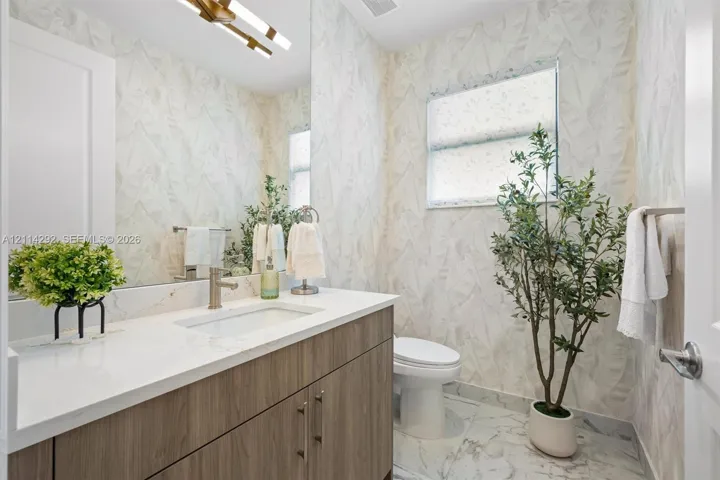 Bathroom featuring a wood-finish vanity with a stone-surface countertop, rectangular undermount sink, patterned wall covering, and polished stone-finish floor tile