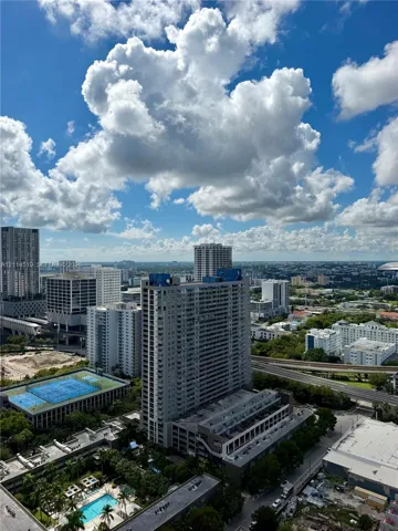 Aerial perspective of urban landscape featuring high-rise architecture, a multi-story parking structure, a blue-surface tennis court, and a rectangular ground-level pool