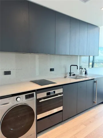 Contemporary kitchen featuring charcoal-toned flat-panel cabinetry, light-toned stone-surface countertops, integrated cooktop, stainless steel wall oven, and light wood-finish flooring