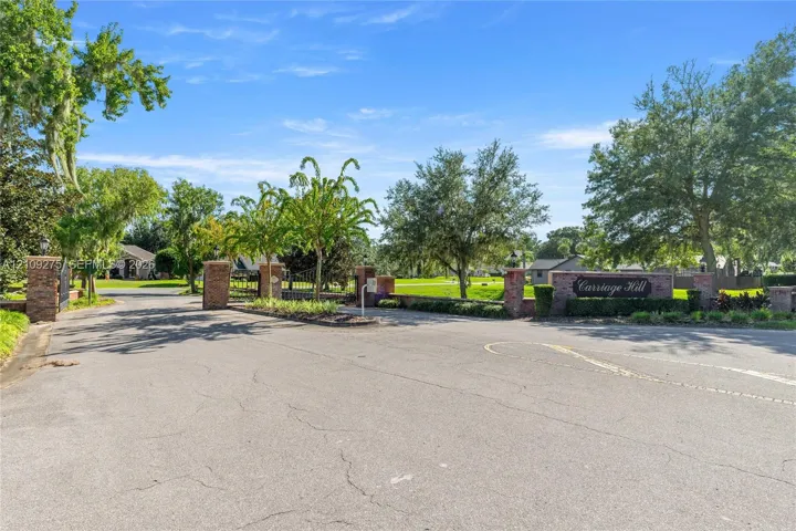 Entryway featuring brick masonry pillars, ornamental metal gate, paved access road, and mature​​‌​​​​‌​​‌‌​‌‌​​​‌‌​‌​‌​‌​​​‌​​ landscaping