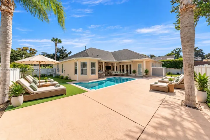 Backyard oasis featuring an in-ground pool, expansive concrete patio, covered lanai, synthetic turf lawn, and mature palm trees