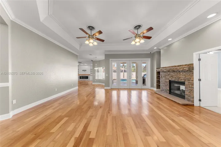 Spacious living area featuring wood-finish flooring, tray ceilings with dual ceiling fans, a stacked-stone fireplace, and multi-panel glass doors
