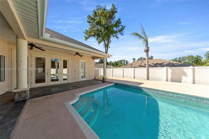 Covered patio with stone-finish flooring and dual ceiling fans, swimming pool with integrated steps, and white vinyl privacy fencing