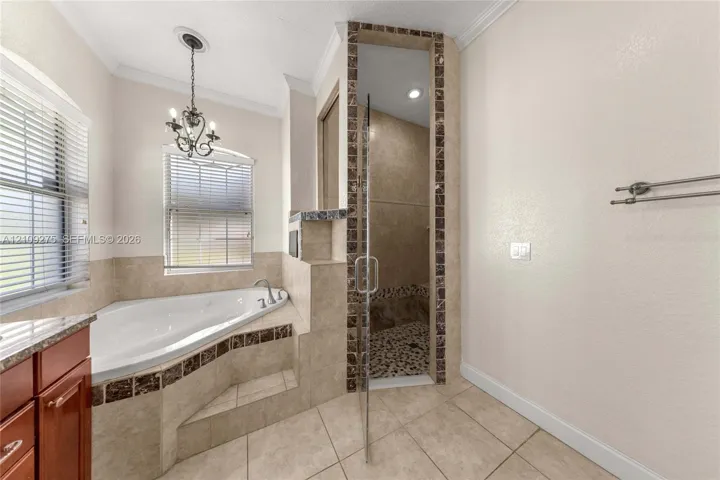 Corner jetted soaking tub featuring decorative tile accents, glass-enclosed shower with stone tile flooring, crown molding, and metal chandelier fixture
