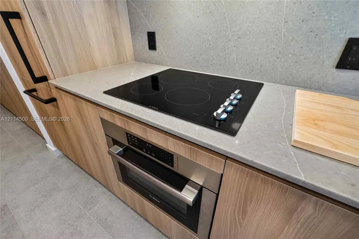 Modern kitchen area featuring flat-panel wood-finish cabinetry, stone-surface countertops, full-height stone backsplash, integrated stainless steel oven, and a black glass electric cooktop