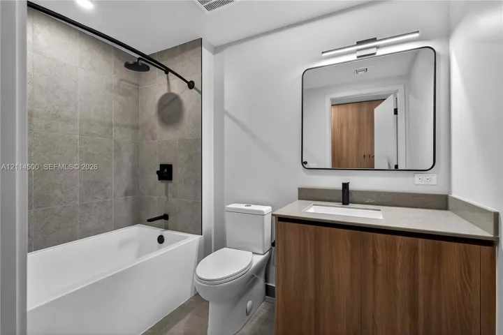 Bathroom featuring a wood-finish vanity with a stone-composite countertop, matte black fixtures, a rectangular undermount sink, and a tiled shower surround