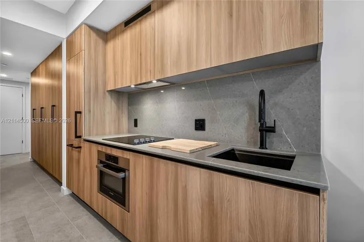 Kitchen featuring wood-finish cabinetry, stone slab backsplash, quartz countertops, undermount sink with matte black faucet, and built-in stainless steel appliances
