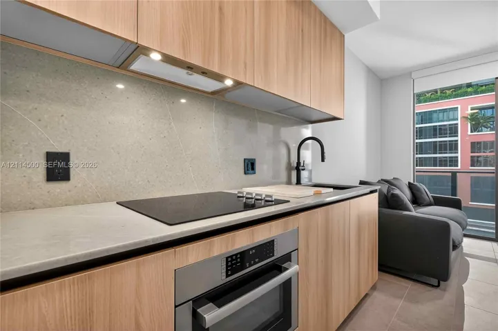 Kitchen featuring light wood-finish cabinetry, stone slab backsplash, integrated induction cooktop, built-in stainless steel oven, and matte black faucet