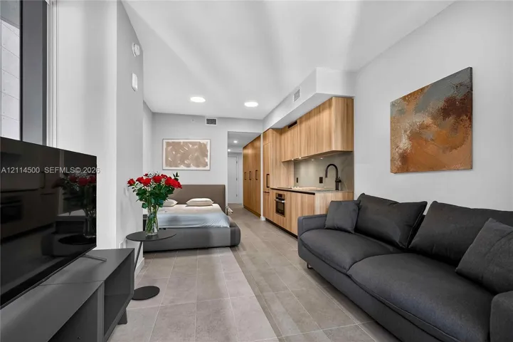 Studio interior featuring wood-finish cabinetry, stone-style tile flooring, recessed ceiling lighting, and a built-in kitchen area with a matte black faucet