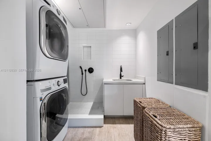 Utility room featuring stackable laundry appliances, a tiled walk-in shower with matte black fixtures, a white vanity with an integrated sink, and recessed lighting