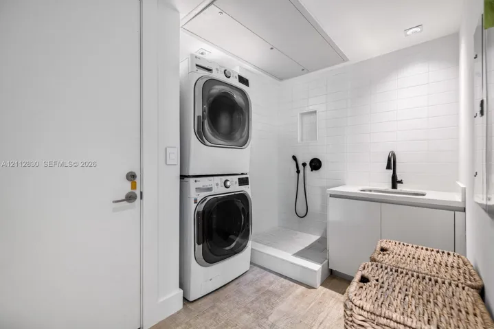 Laundry area featuring stacked appliances, tiled shower enclosure with handheld sprayer, built-in cabinetry, solid-surface countertop, and matte black faucet