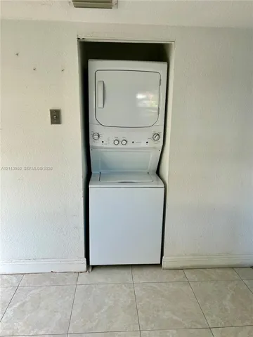 Stacked white laundry unit set within a recessed wall alcove, featuring a top-mounted dryer, bottom-loading washer, and neutral ceramic tile flooring