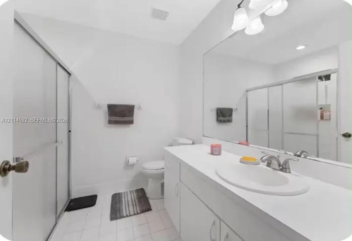 Full-width mirror, white cabinetry with solid-surface countertop, integrated vanity sink, brushed-metal faucet, and walk-in shower enclosure