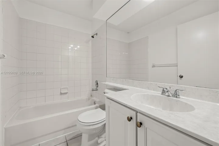 Bathroom featuring a white bathtub with a tile surround, vanity with a light-toned stone-patterned countertop, integrated oval sink, and a full-width wall mirror
